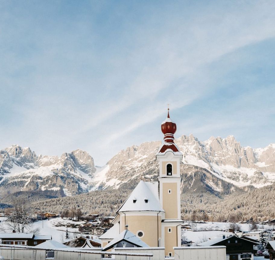 Verschneites Dorf mit Kirche im Vordergrund, umgeben von den schneebedeckten Bergen des Wilden Kaisers unter blauem Himmel.