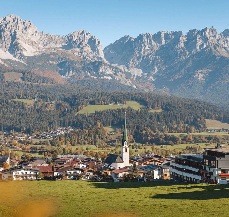 Panoramic view of the Wilder Kaiser mountains above a Tyrolean village, featuring green fields, a church spire, and majestic peaks under a blue sky.
