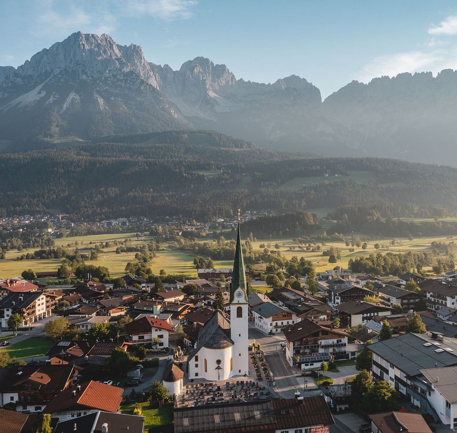 A charming village with a prominent church spire lies under the impressive Wilder Kaiser mountain range, illuminated by the gentle morning light.
