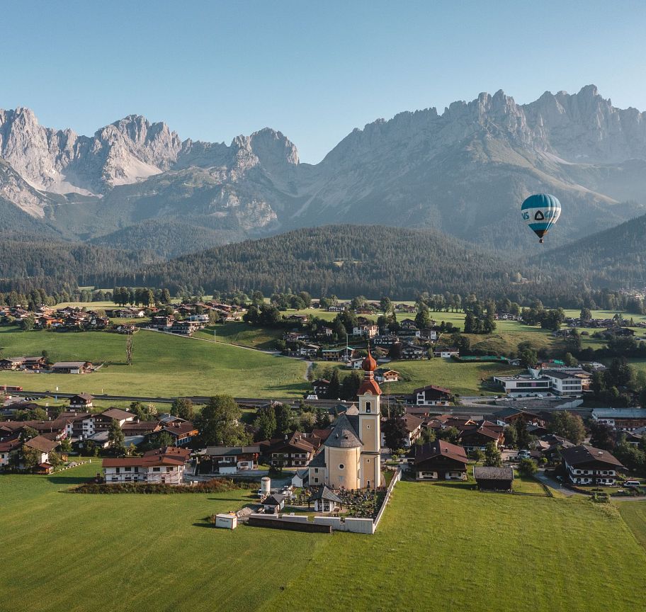 Scenic view of the Wilder Kaiser mountains with a hot air balloon over a village and green fields.