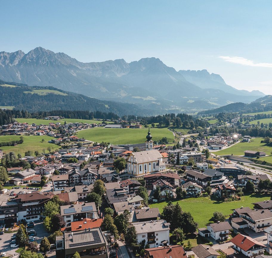 Luftaufnahme eines malerischen Dorfes in Tirol mit alpiner Kulisse, umgeben von Wiesen und Bergen. Im Hintergrund ragt das imposante Wilder Kaiser Massiv empor.