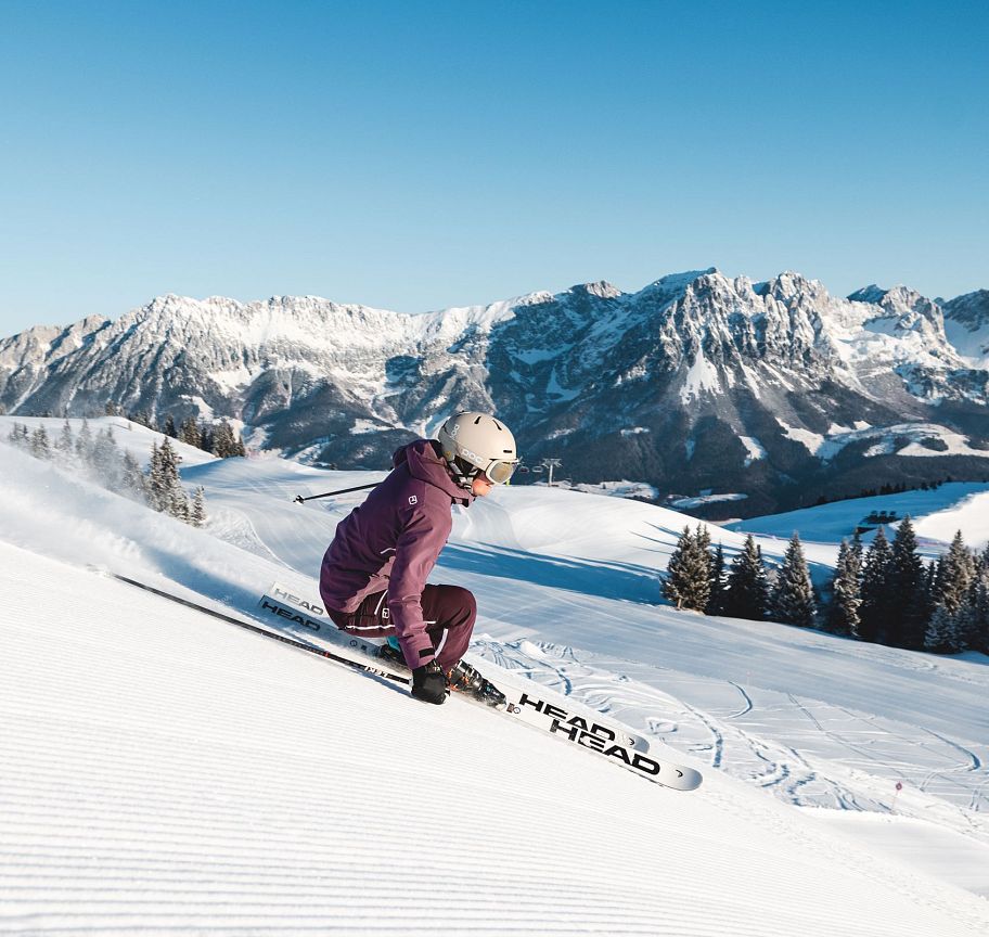 Skier in a purple jacket descends snowy slopes at Wilder Kaiser with mountain range in the background under a clear blue sky.