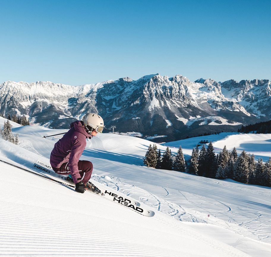 Eine Person fährt auf Skiern eine schneebedeckte Piste hinunter, umgeben von einem beeindruckenden Bergpanorama unter klarem, blauem Himmel.