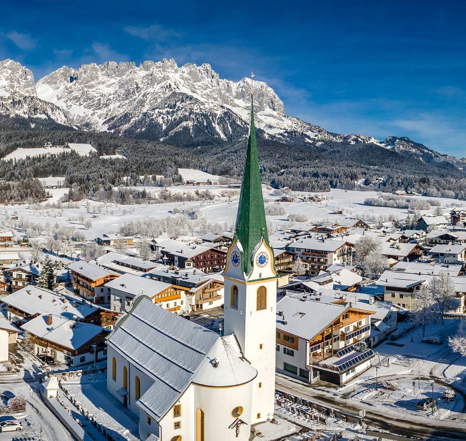 Snow-covered village with a church tower in foreground, set against a backdrop of towering mountains under a clear blue sky at Wilder Kaiser, Austria.