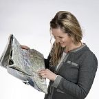 A woman smiles as she looks at a large unfolded map, indicating a spot with her finger. She's wearing a gray jacket and has long, wavy hair.