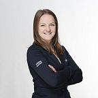 Eine lächelnde Person mit braunem Haar, gekleidet in eine dunkle Jacke mit der Aufschrift "Wilder Kaiser", die vor einem schlichten Hintergrund posiert.