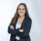 Junge Frau mit langen Haaren und Brille lächelt in die Kamera. Sie trägt einen dunklen Blazer, eine helle Bluse und eine Perlenkette. Hintergrund ist neutral.