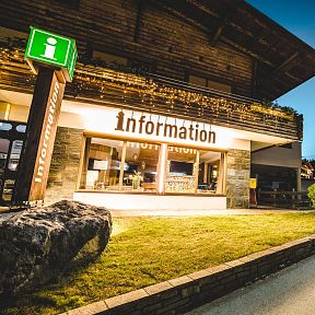 Ein beleuchtetes Informationszentrum in einem alpinen Dorf bei Dämmerung, mit einem Kirchturm im Hintergrund und einem Schild mit dem Buchstaben "i".
