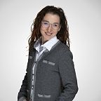 Person mit lockigem Haar, Brille und grauem Blazer lächelt freundlich vor weißem Hintergrund.