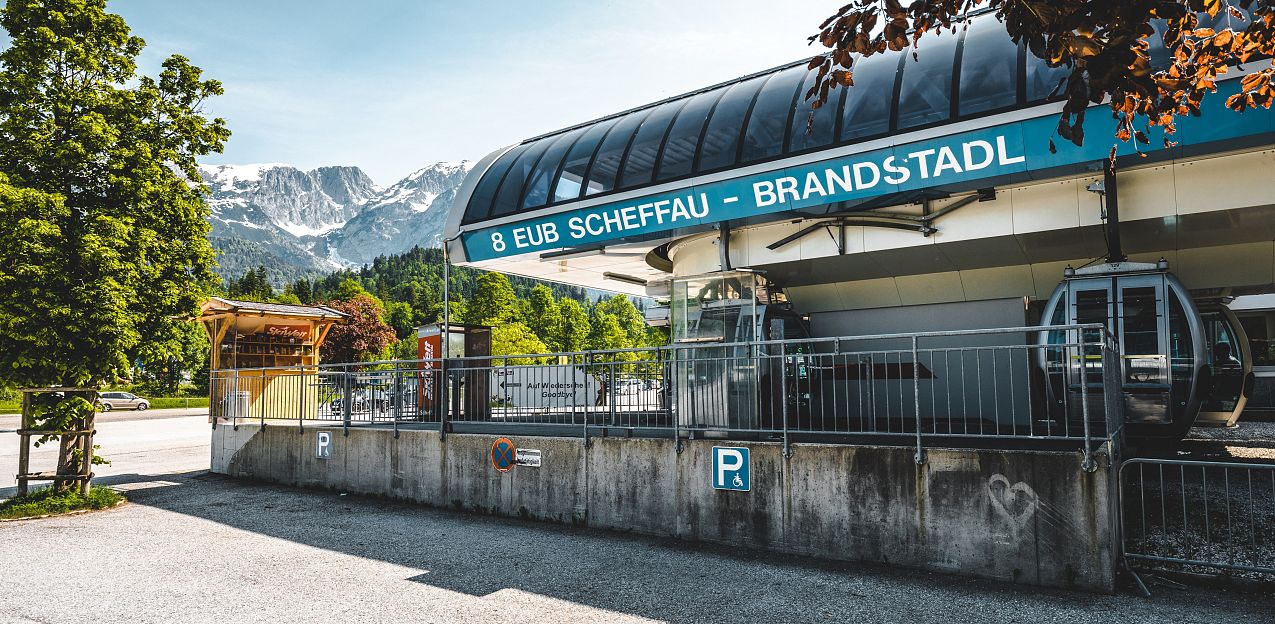 Seilbahnstation in Scheffau, Tirol, mit grüner Landschaft und beeindruckendem Bergmassiv im Hintergrund unter klarem Himmel.