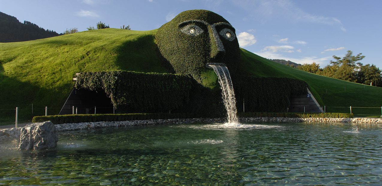 Künstlerischer Rasenhügel mit Gesichtsskulptur und Wasserfall als Mund, umgeben von einem flachen Teich unter blauem Himmel, in sonniger Landschaft.