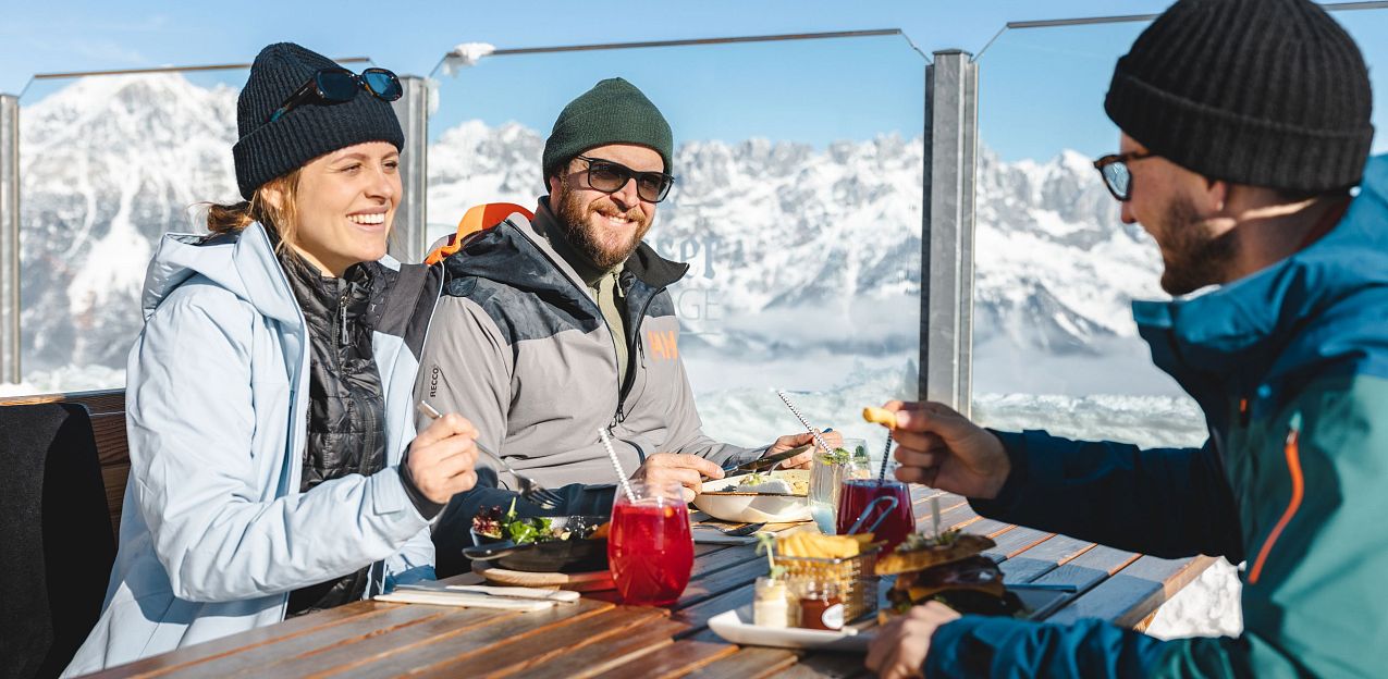 Three people dining outdoors at a wooden table, with snow-covered mountains of the Wilder Kaiser visible in the background, under a clear blue sky.