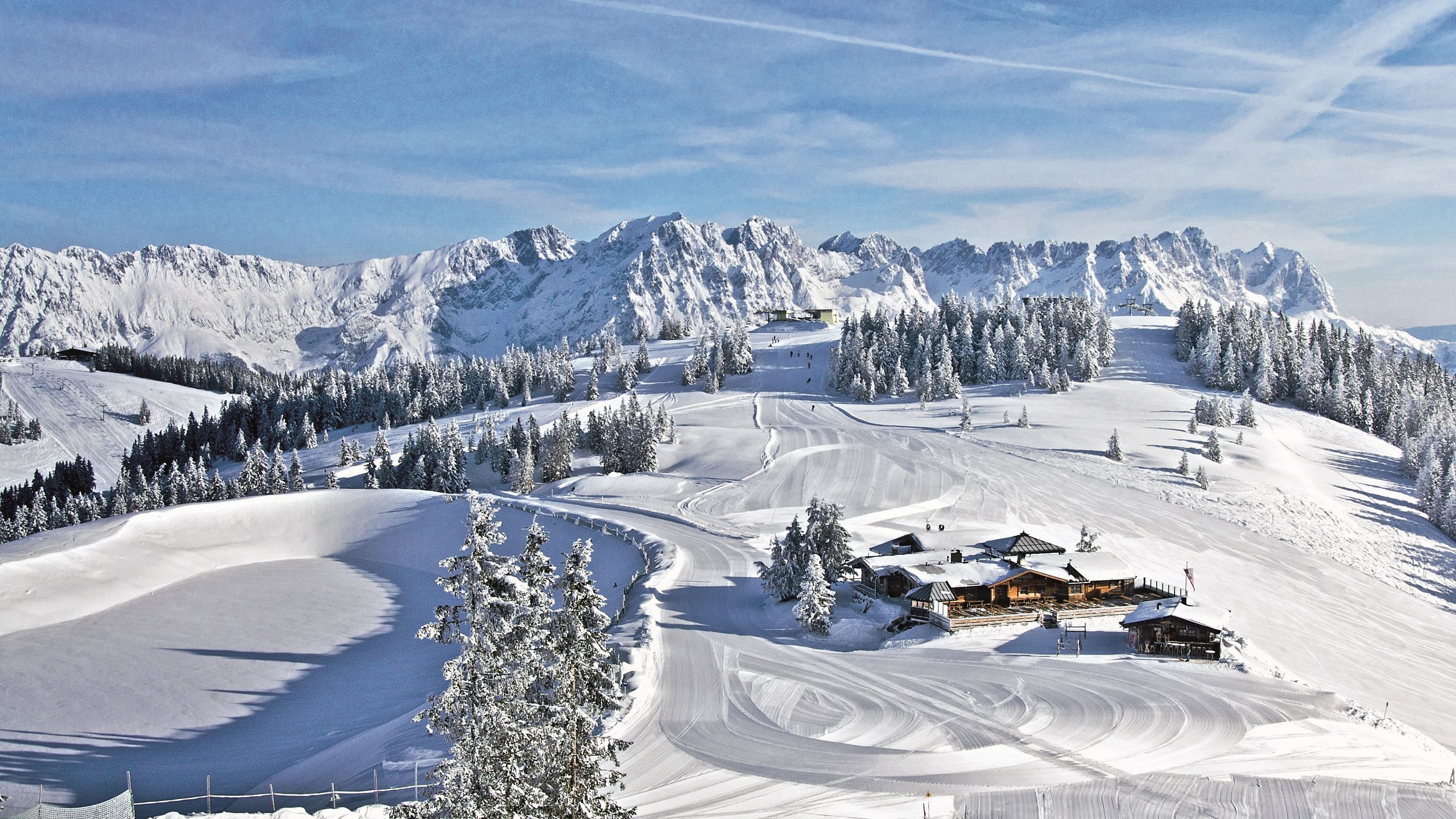 Verschneite Berglandschaft in Tirol mit einem kleinen Chalet in der Mitte, umgeben von Bäumen und dem beeindruckenden Wilden Kaiser im Hintergrund.