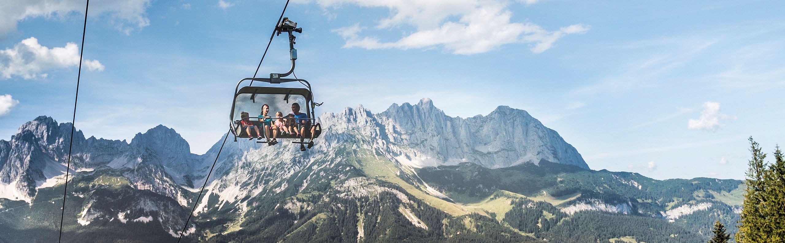 Scenic view of the Wilder Kaiser mountain range with a cable car in the foreground, surrounded by lush greenery and a clear blue sky.