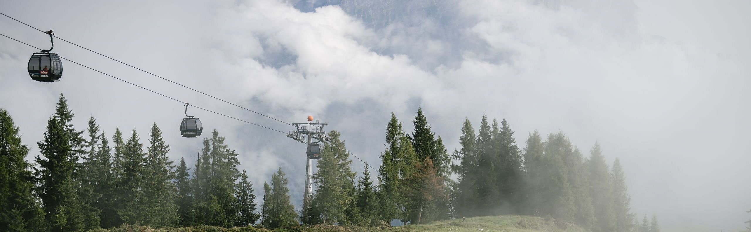 Malerische Berglandschaft mit Seilbahn, umgeben von dichtem Nebel und grünen Wiesen, im Hintergrund majestätische Gipfel. Verträumte Atmosphäre in den Alpen.