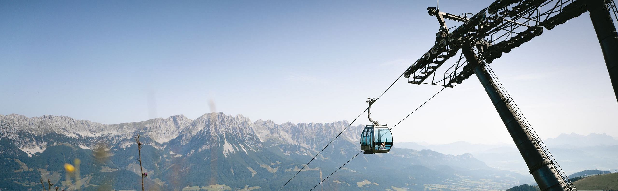 A cable car ascends a green hillside with a majestic mountain range in the background under a clear blue sky.