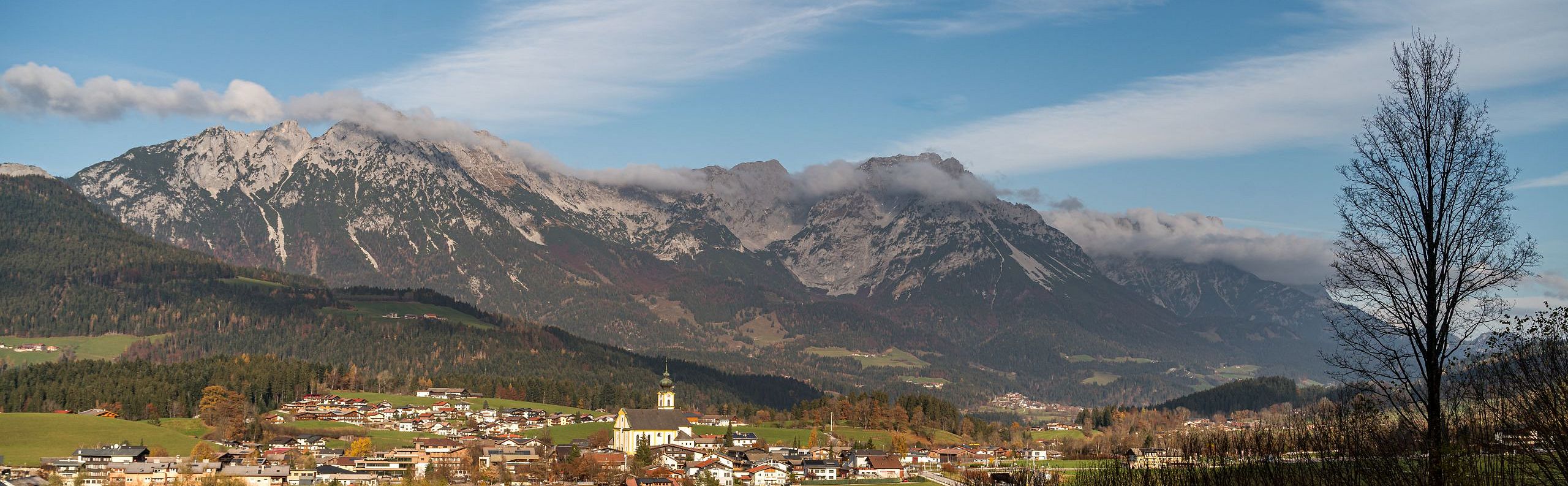Panoramablick auf das Tiroler Bergmassiv Wilder Kaiser mit einer idyllischen Ortschaft im Vordergrund, umgeben von grünen Wiesen und bewaldeten Hügeln.