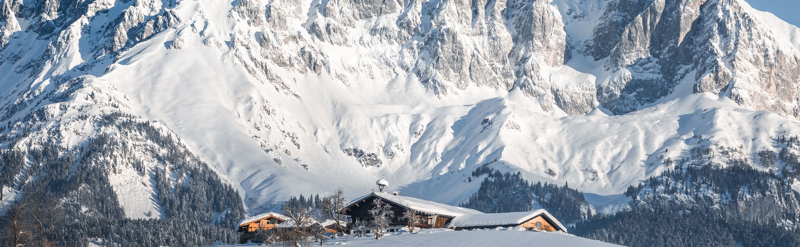 Snow-covered alpine landscape with wooden huts and a dramatic mountain range in the background under a clear blue sky.