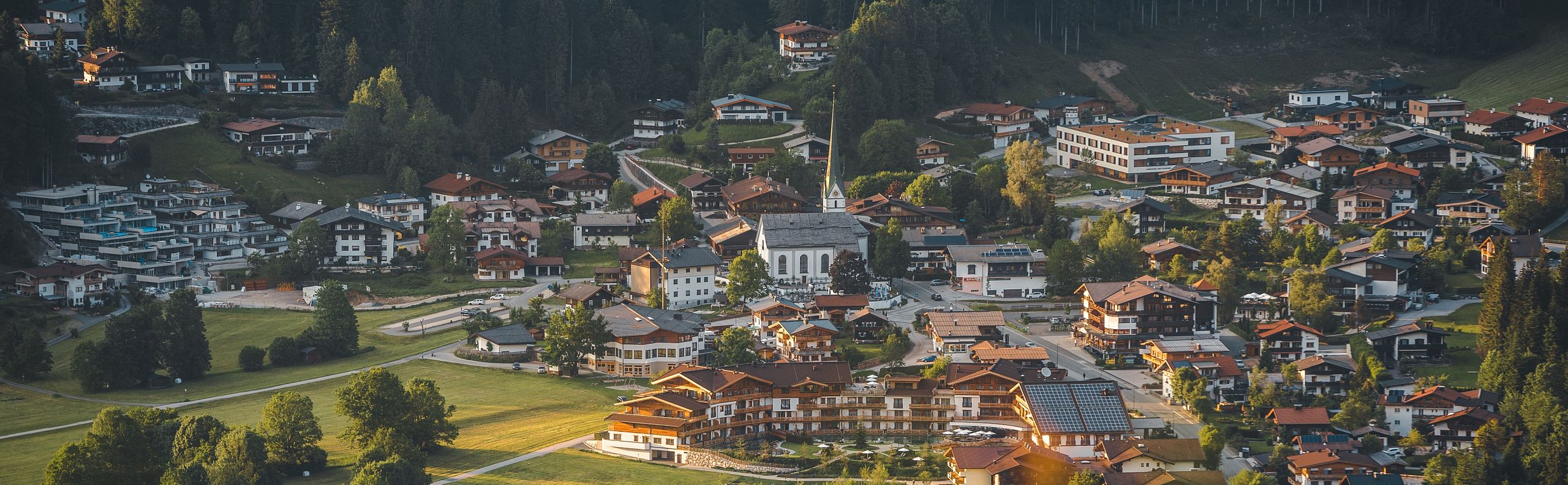 Luftaufnahme eines malerischen Dorfes in einer grünen Landschaft, umgeben von Wäldern und Hügeln, mit einer zentral gelegenen Kirche und verstreuten Häusern.