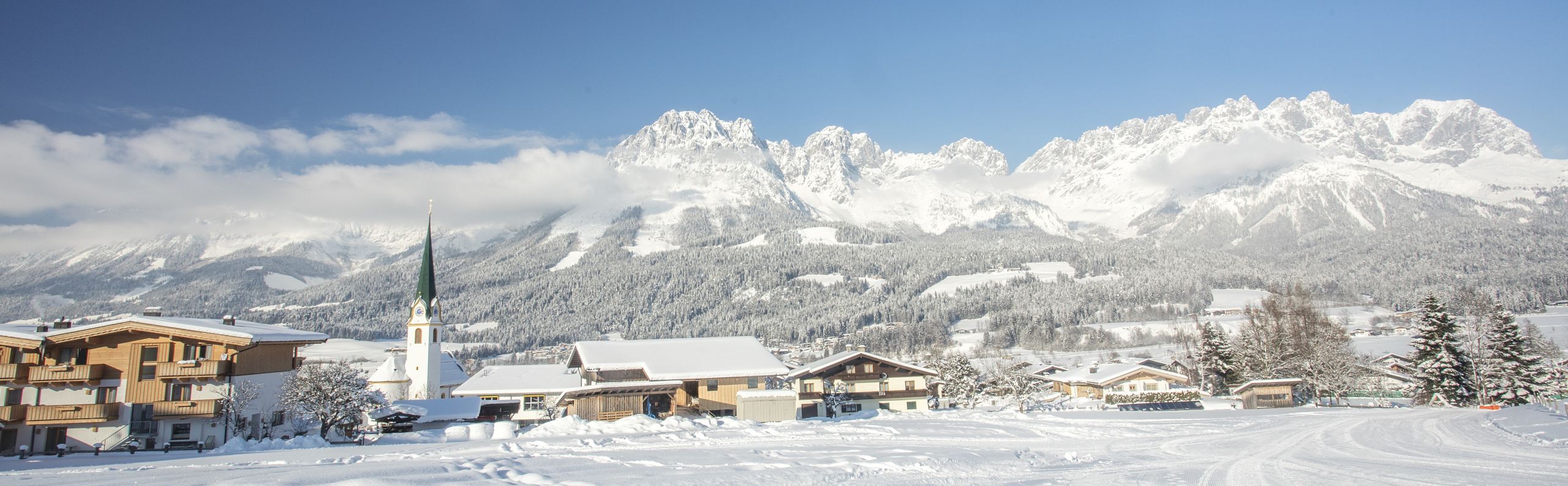 A snow-covered village with a church and houses set against the backdrop of the Wilder Kaiser mountains under a bright blue sky.