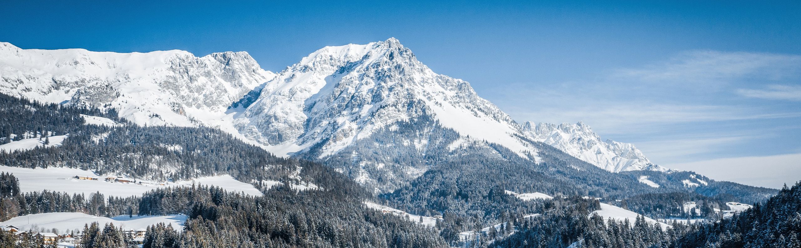 Snow-covered village with the majestic Wilder Kaiser mountains in the background, under a clear blue sky, capturing the essence of a peaceful winter day.
