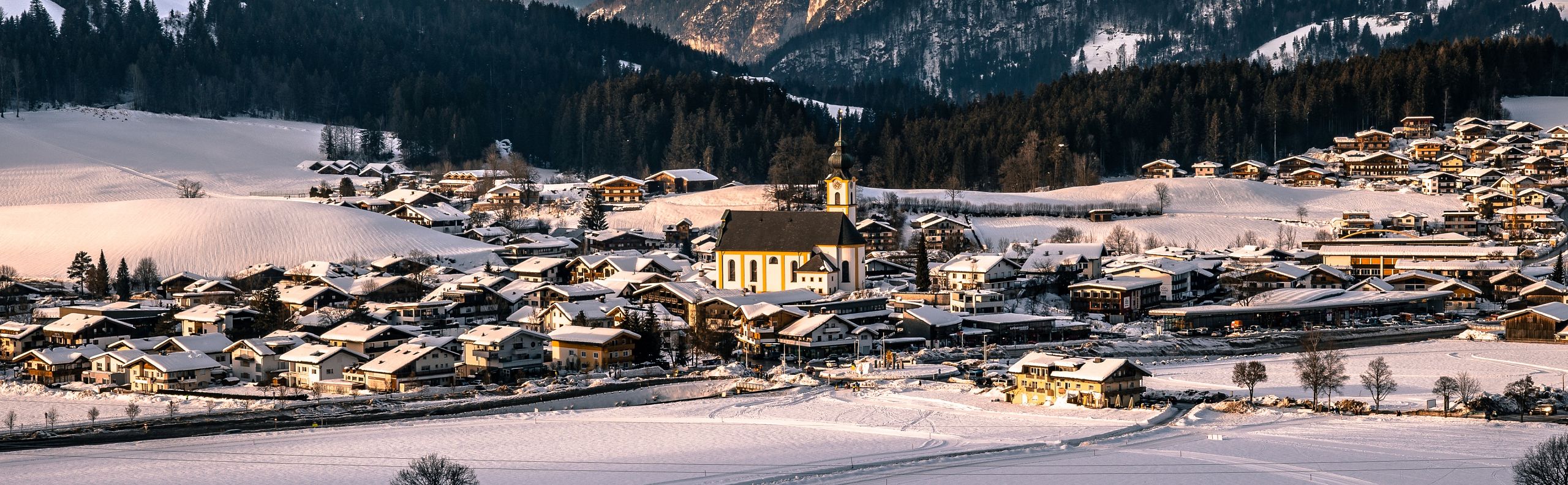 A snow-covered village nestled in the Wilder Kaiser region, with mountains in the background and warm sunlight illuminating the landscape.