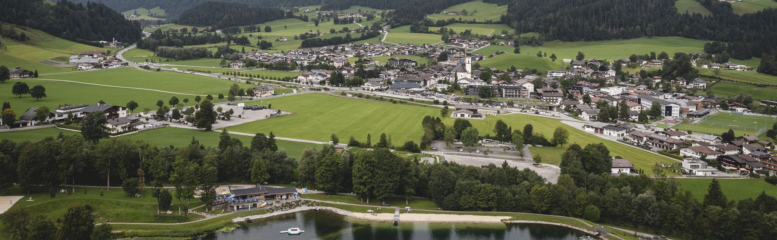 Panoramablick auf eine grüne Berglandschaft mit einem kleinen See im Vordergrund, umgeben von Wäldern und einem Dorf am Fuße imposanter Berge.