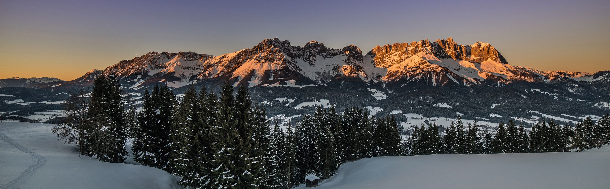 Snow-covered mountains at sunset with a clear sky and forest in foreground, creating a serene winter landscape.
