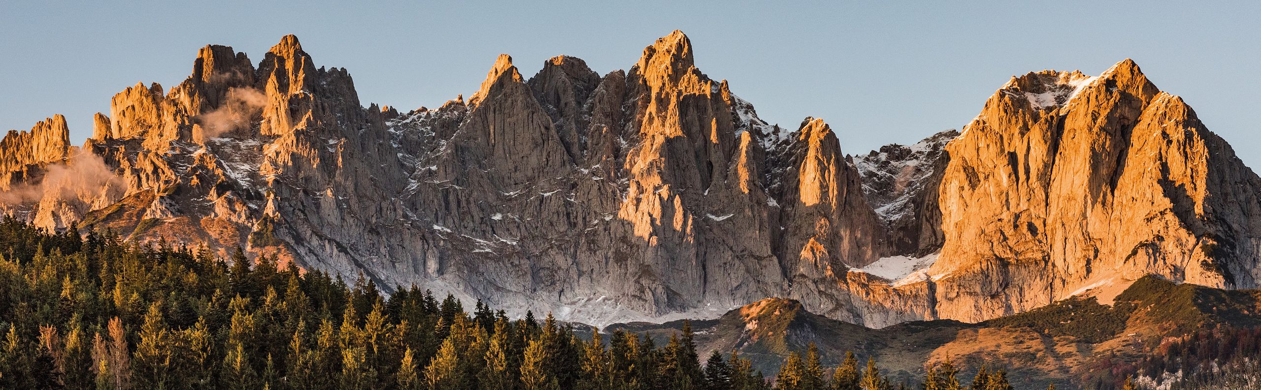 Beeindruckendes Bergmassiv des Wilden Kaisers bei Sonnenuntergang, mit Felsen in warmem Licht und einem Wald im Vordergrund.