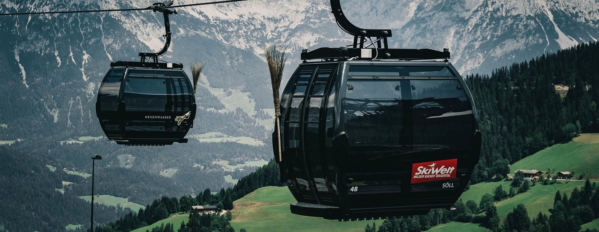Zwei moderne Gondeln schweben über grüne Hänge vor einer beeindruckenden Bergkulisse in den Alpen, mit klarem Himmel und vereinzelten Wolken.