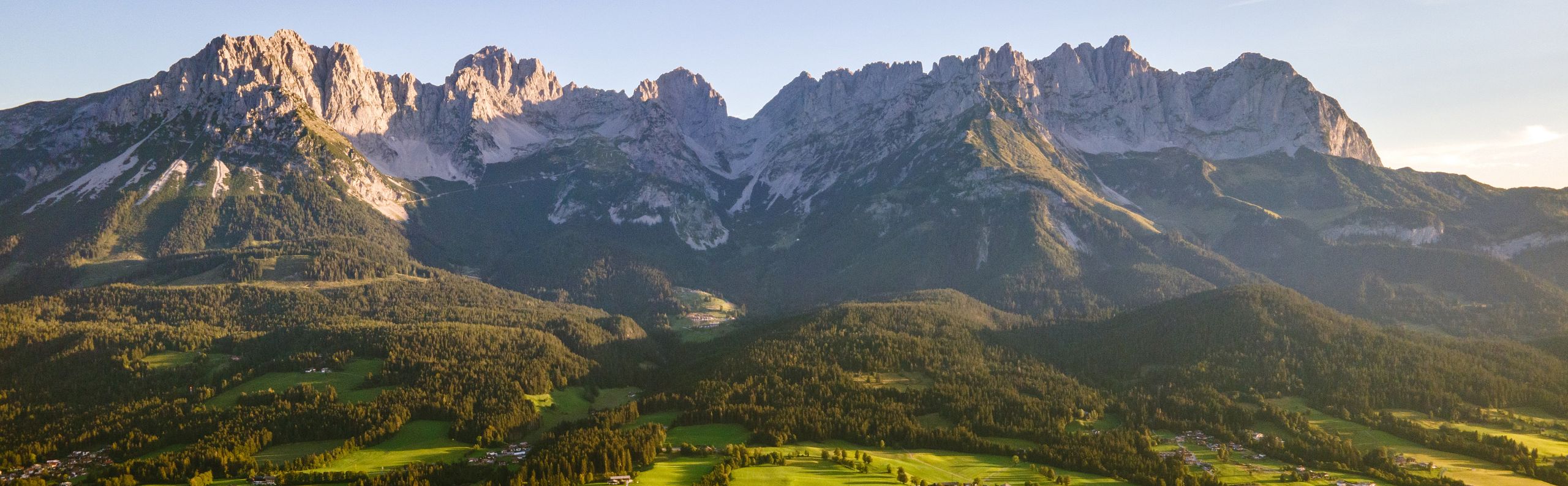 Panoramablick auf das imposante Bergmassiv des Wilden Kaisers in Tirol, eingerahmt von grünen Wiesen und malerischen Dörfern unter einem klaren blauen Himmel.