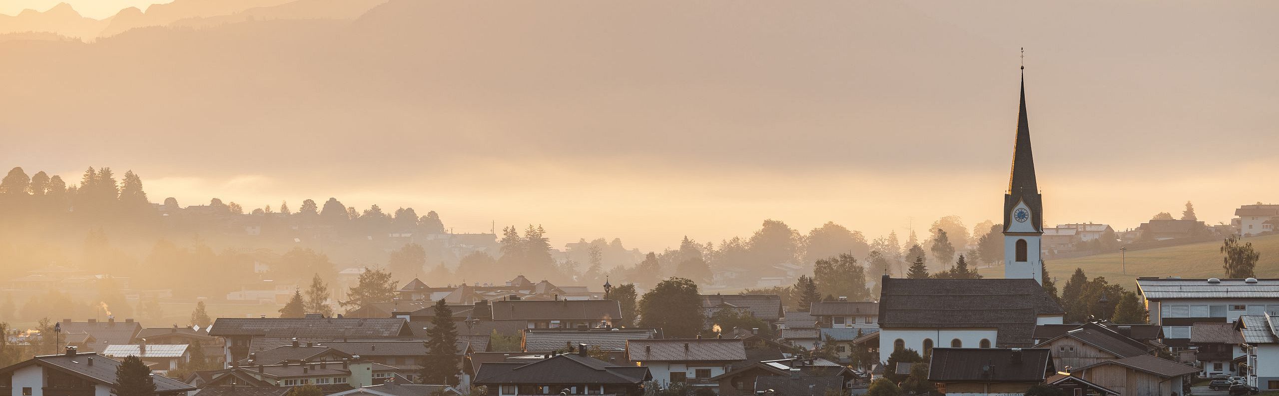 Bergdorf bei Sonnenaufgang mit Kirche im Vordergrund und majestätischen Bergen im Hintergrund, umhüllt von Morgennebel.