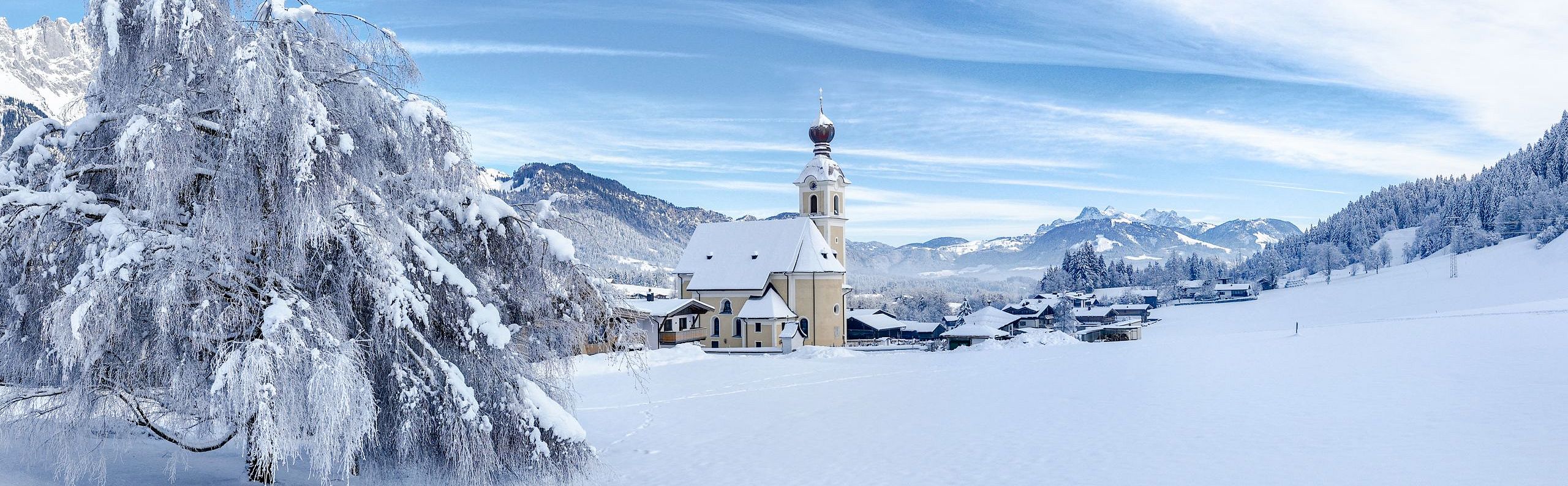 Verschneite Landschaft mit einer Kirche im Hintergrund, umgeben von Bergen und einem beeindruckend blauen Himmel mit zarten Wolkenstreifen.