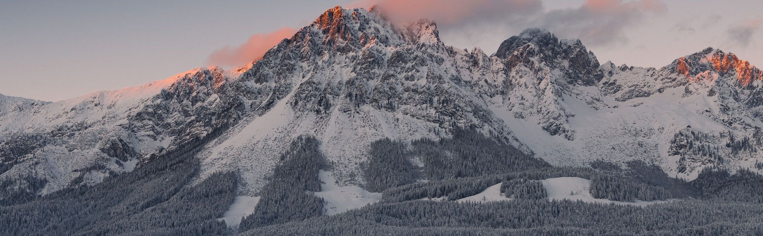Mountain peaks of the Wilder Kaiser glowing with sunrise hues, above snowy forests and clouds. A serene scene capturing nature's beauty at dawn.