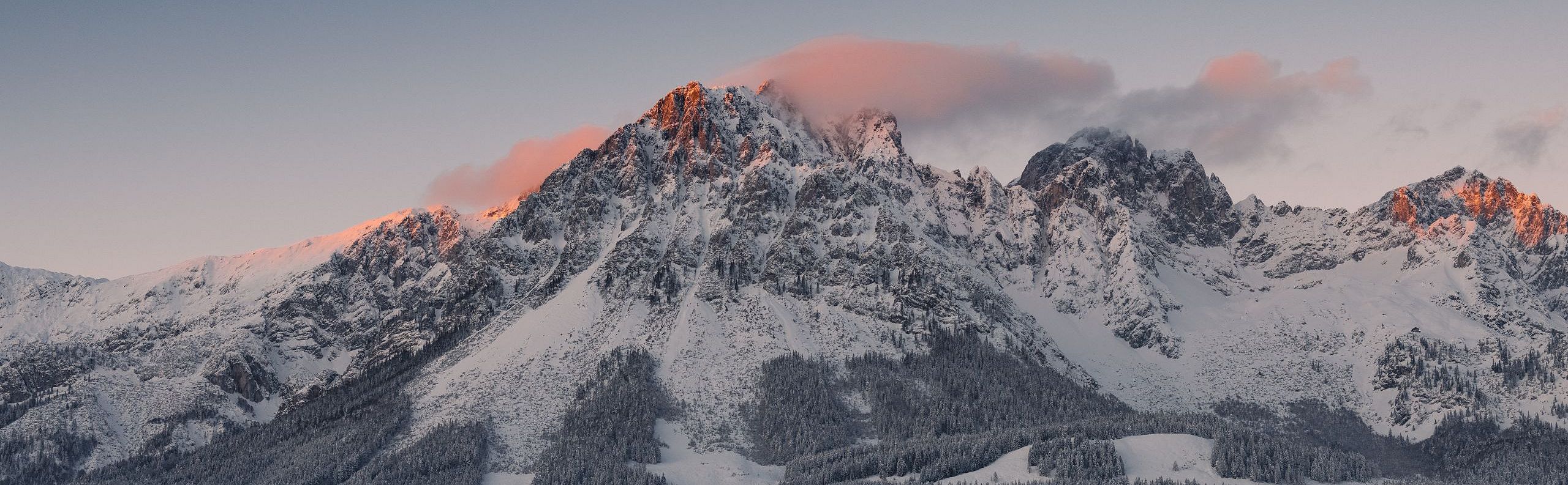 Mountain peaks of the Wilder Kaiser glowing with sunrise hues, above snowy forests and clouds. A serene scene capturing nature's beauty at dawn.