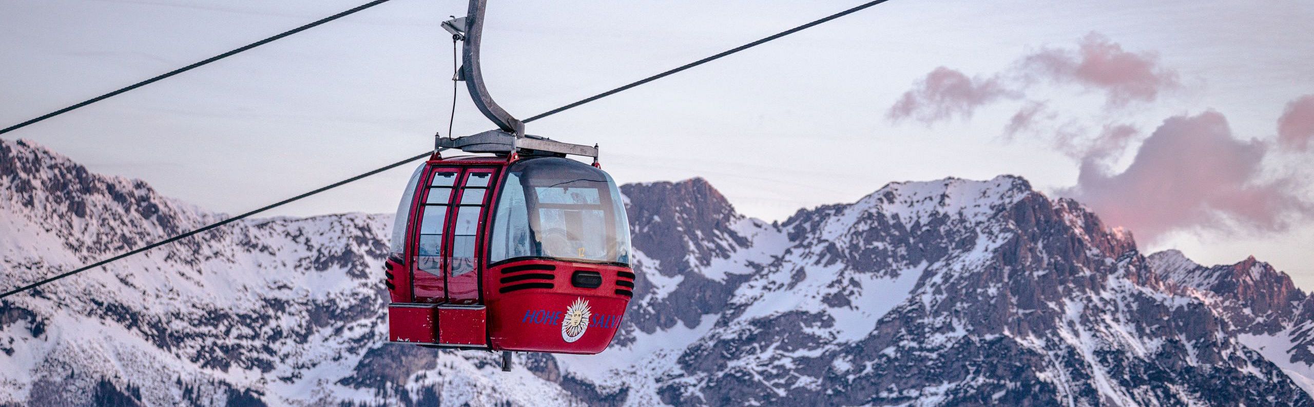 Rote Seilbahngondel vor schneebedeckten Berggipfeln bei Sonnenuntergang, mit pastellfarbenem Himmel im Hintergrund. Winterliche Landschaft in den Alpen.