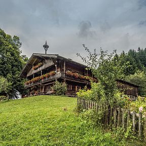 Traditionelles Tiroler Bauernhaus mit Holzfassade und Balkonblumen vor einem grünen Hang, umgeben von Bäumen und einem Holzzaun, unter bewölktem Himmel.