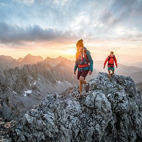Zwei Wanderer in sportlicher Kleidung erklimmen einen felsigen Berggipfel bei Sonnenuntergang, mit beeindruckender Bergkulisse im Hintergrund.