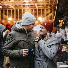 A couple in winter clothing enjoy hot drinks at a festive outdoor market, surrounded by people and lights, with gentle snowfall in the background.