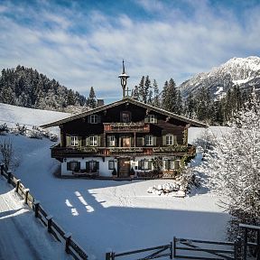 Traditionelles Tiroler Holzhaus im Winter, umgeben von schneebedeckten Bäumen und Bergen im Hintergrund. Die Landschaft ist idyllisch und ruhig.