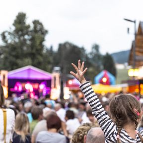 Outdoor concert scene with a large crowd and a young girl with raised arms enjoying the music. Colorful stage lights and tents add to the festive atmosphere.