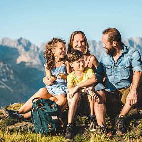 Eine Familie sitzt lachend auf einem Bergwiese mit Rucksack, im Hintergrund ein beeindruckendes Bergpanorama unter einem klaren blauen Himmel.