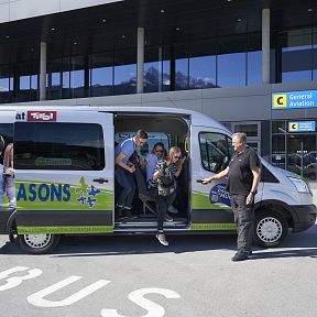 A shuttle bus parked at a terminal, with passengers exiting, ready to explore the Wilder Kaiser region, in front of a modern building.