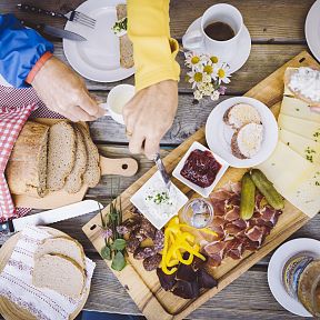 Gemütliches Picknick auf Holztisch mit Brot, Käse, Aufschnitt, eingelegtem Gemüse und Getränken. Zwei Hände greifen nach Essen, rustikale Atmosphäre.