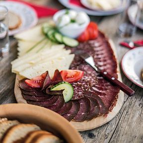 Eine rustikale Jause mit Käse, Wurst, Tomaten, Gurken und Brot auf einem Holztisch, appetitlich angerichtet. Perfekt für ein traditionelles Tiroler Mahl.