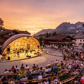Open-Air-Konzert in idyllischer Bergdorfkulisse bei Sonnenuntergang. Eine Bühne mit Publikum, umgeben von traditionellen Gebäuden und majestätischen Bergen.