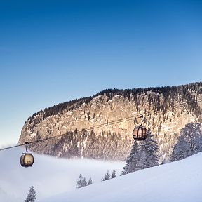 Zwei schwebende Gondeln vor einer verschneiten Bergkulisse unter klarem, blauem Himmel. Dichte Nebelfelder liegen im Tal, flankiert von schneebedeckten Bäumen.