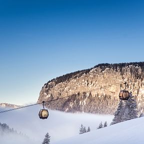 Zwei schwebende Gondeln vor einer verschneiten Bergkulisse unter klarem, blauem Himmel. Dichte Nebelfelder liegen im Tal, flankiert von schneebedeckten Bäumen.