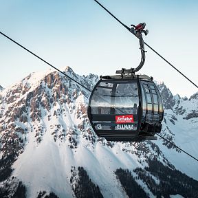 Eine Gondelbahn schwebt über schneebedeckten Bergspitzen im Skigebiet Ellmau, Wilder Kaiser, vor strahlend blauem Himmel im Winter.