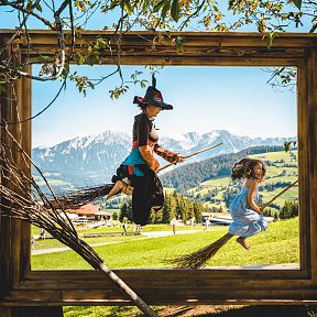 Zwei Personen in Kostümen fliegen auf Besen in einem großen Holzrahmen, mit Bergen und grüner Landschaft im Hintergrund, unter einem Baum befestigt.